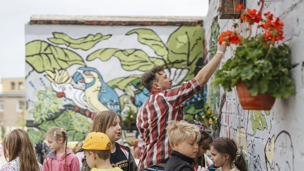 Grupa dzieci i dorosły wspólnie malują mural na ścianie, wokół rosną kwiaty, a tło muralu stanowią duże liście i kolorowe ptaki, scena utrzymana w atmosferze twórczej współpracy na świeżym powietrzu.