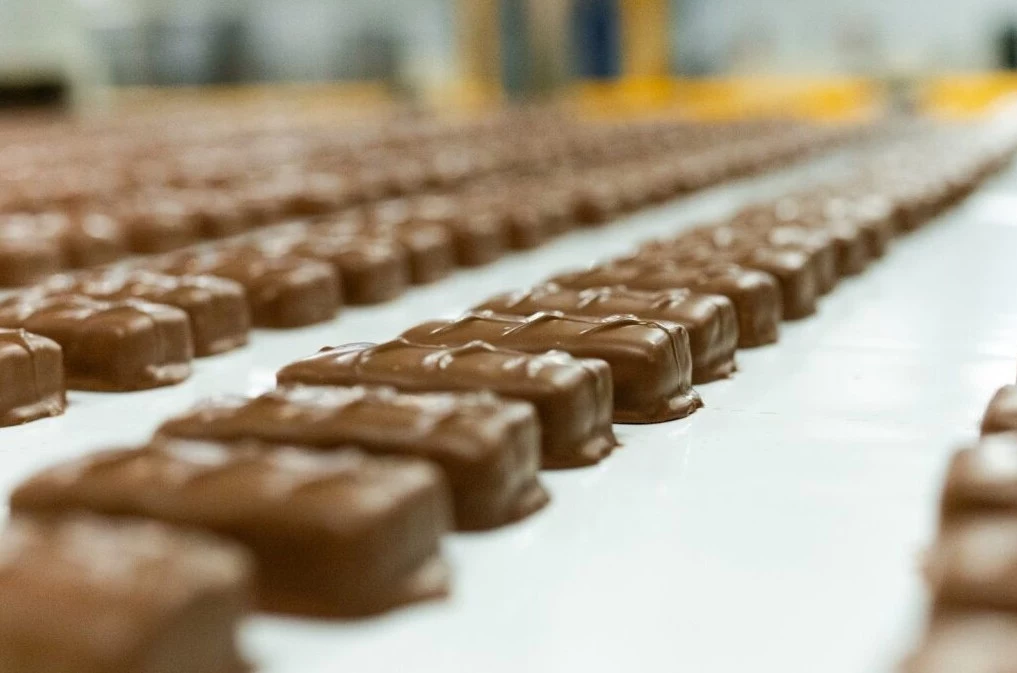 Rzędy czekoladowych pralinek ustawione na taśmie produkcyjnej w fabryce, każda z nich pokryta gładką warstwą mlecznej czekolady, w tle rozmyte elementy hali produkcyjnej.