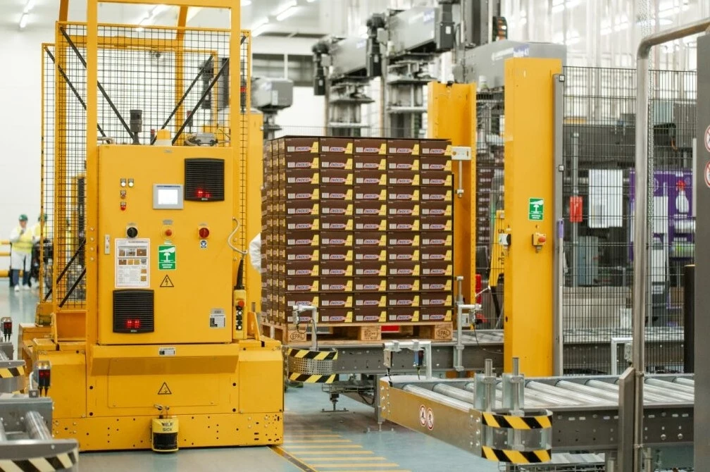 Automatyczna linia produkcyjna z żółtymi maszynami przemysłowymi i zapakowanymi na palecie opakowaniami produktów gotowych, z metalowymi barierkami ochronnymi i przenośnikami taśmowymi.