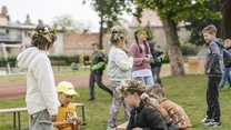 Dzieci bawiące się przy drewnianych skrzynkach z naturalnymi materiałami podczas zajęć edukacyjnych na świeżym powietrzu, niektóre noszą wianki z kwiatów; w tle park i ławki.
