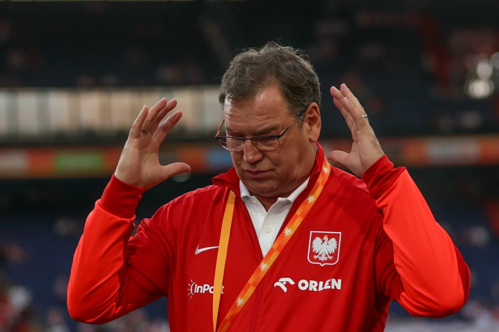 Mężczyzna w czerwonej kurtce reprezentacji Polski z herbem orzełka na piersi, trzyma ręce przy głowie w geście rozczarowania lub skupienia, w tle rozmyte trybuny stadionu.