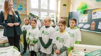 Grupa dzieci ubranych w białe koszulki z logo "Czyste Zielone Miasta" stoi razem w klasie, obok nich nauczycielka. W tle widać balony, plakaty edukacyjne związane z ekologią oraz stół z materiałami do zajęć.