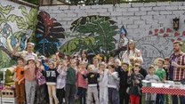 Grupa dzieci i kilku dorosłych stoi razem na tle kolorowej, muralowej ściany z motywami roślinnymi i bajkowymi postaciami, dzieci uśmiechają się i pozują do zdjęcia, po prawej stronie widać stół z materiałami warsztatowymi.