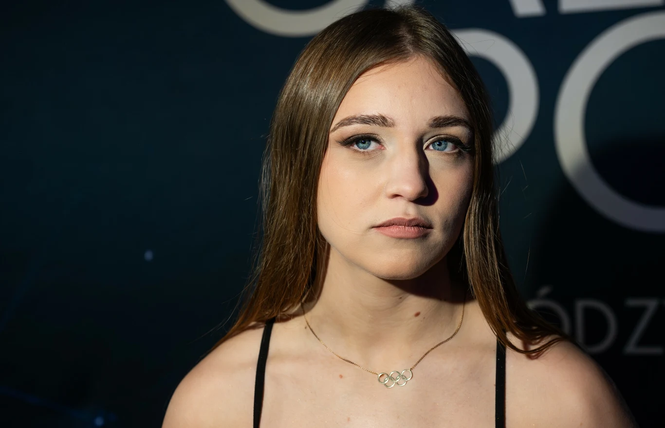 Julia Szeremeta Młoda kobieta o długich, prostych włosach, patrząca przed siebie z neutralnym wyrazem twarzy, nosi delikatny naszyjnik i czarny top na ramiączkach, w tle ciemna, rozmyta grafika.