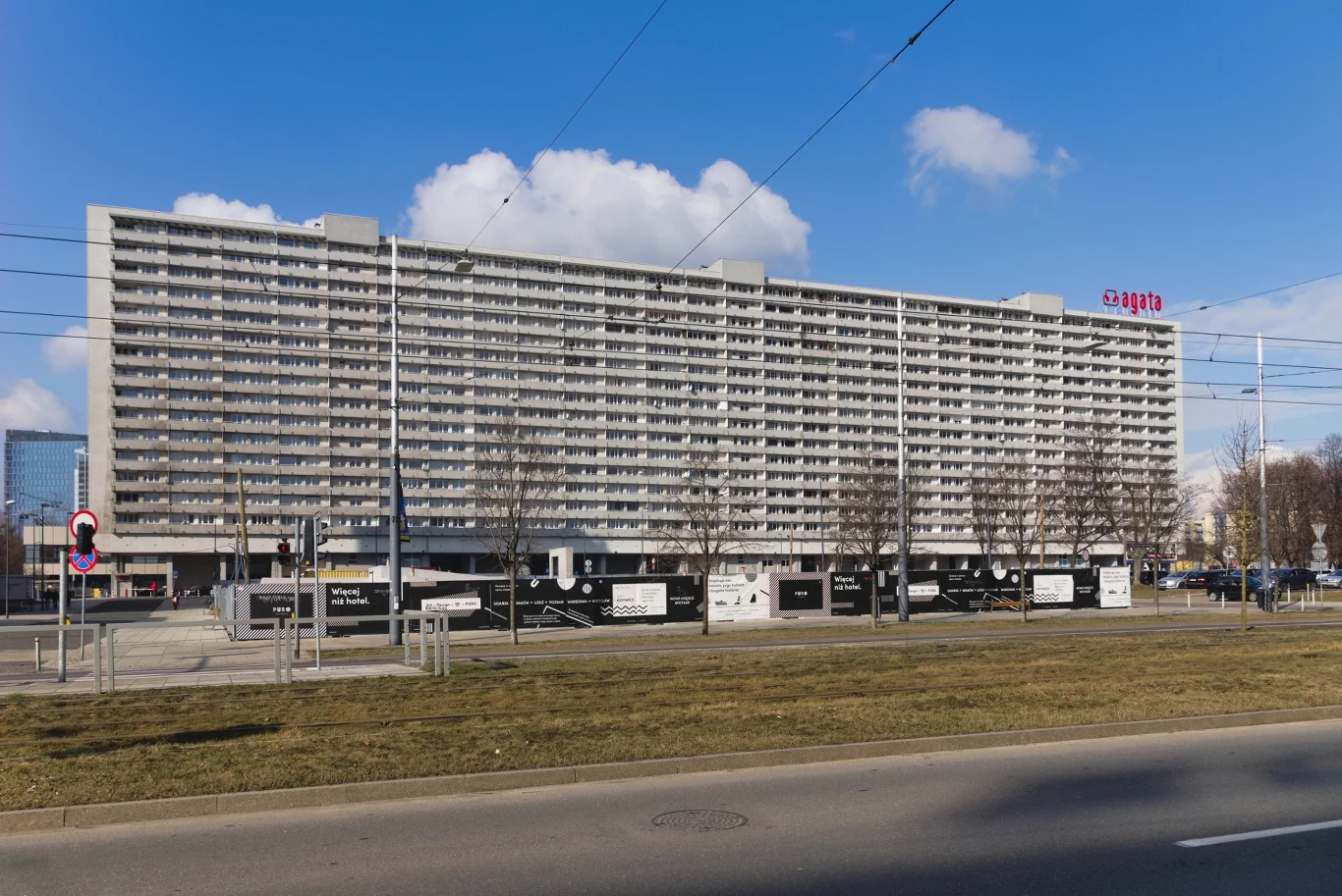 Wielopiętrowy blok mieszkalny o nowoczesnej architekturze, z licznymi balkonami, położony przy szerokiej ulicy. Przed budynkiem znajdują się drzewa o bezlistnych gałęziach i reklamowe tablice ogrodzeniowe.