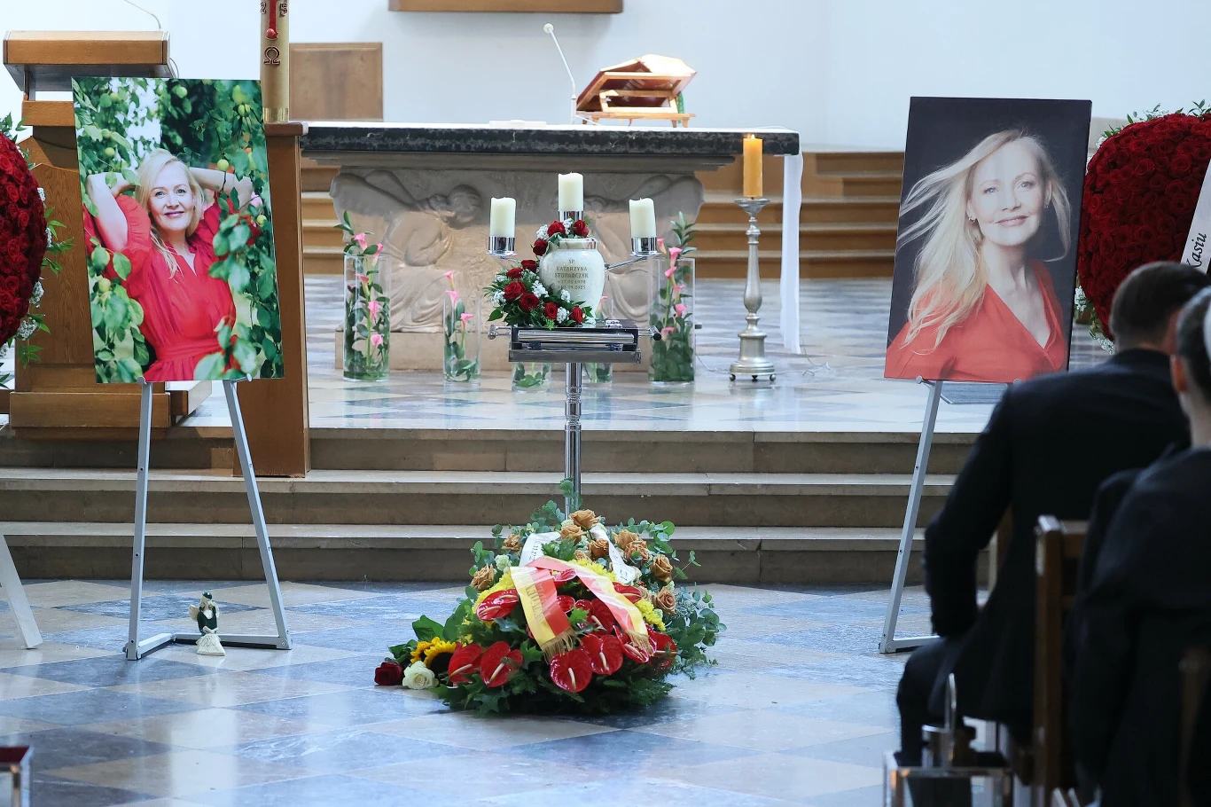 Ceremonia pogrzebowa w kościele, urna z wieńcem kwiatowym ustawiona na piedestale przed ołtarzem, po bokach dwa duże portrety kobiety w czerwonej sukience oraz liczne kwiaty i świece.