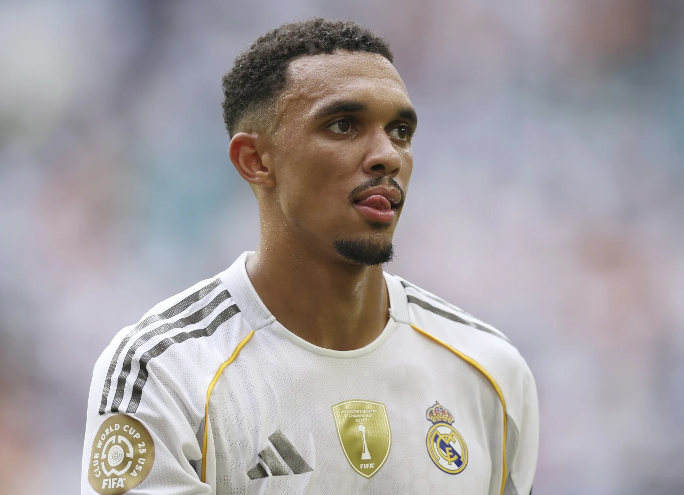 Trent Alexander-Arnold Piłkarz w białym stroju Realu Madryt z herbami klubowymi i odznaką mistrzowską FIFA na rękawie, stoi na tle rozmytego stadionu, patrząc przed siebie z poważnym wyrazem twarzy, z widocznym wysiłkiem sportowym.