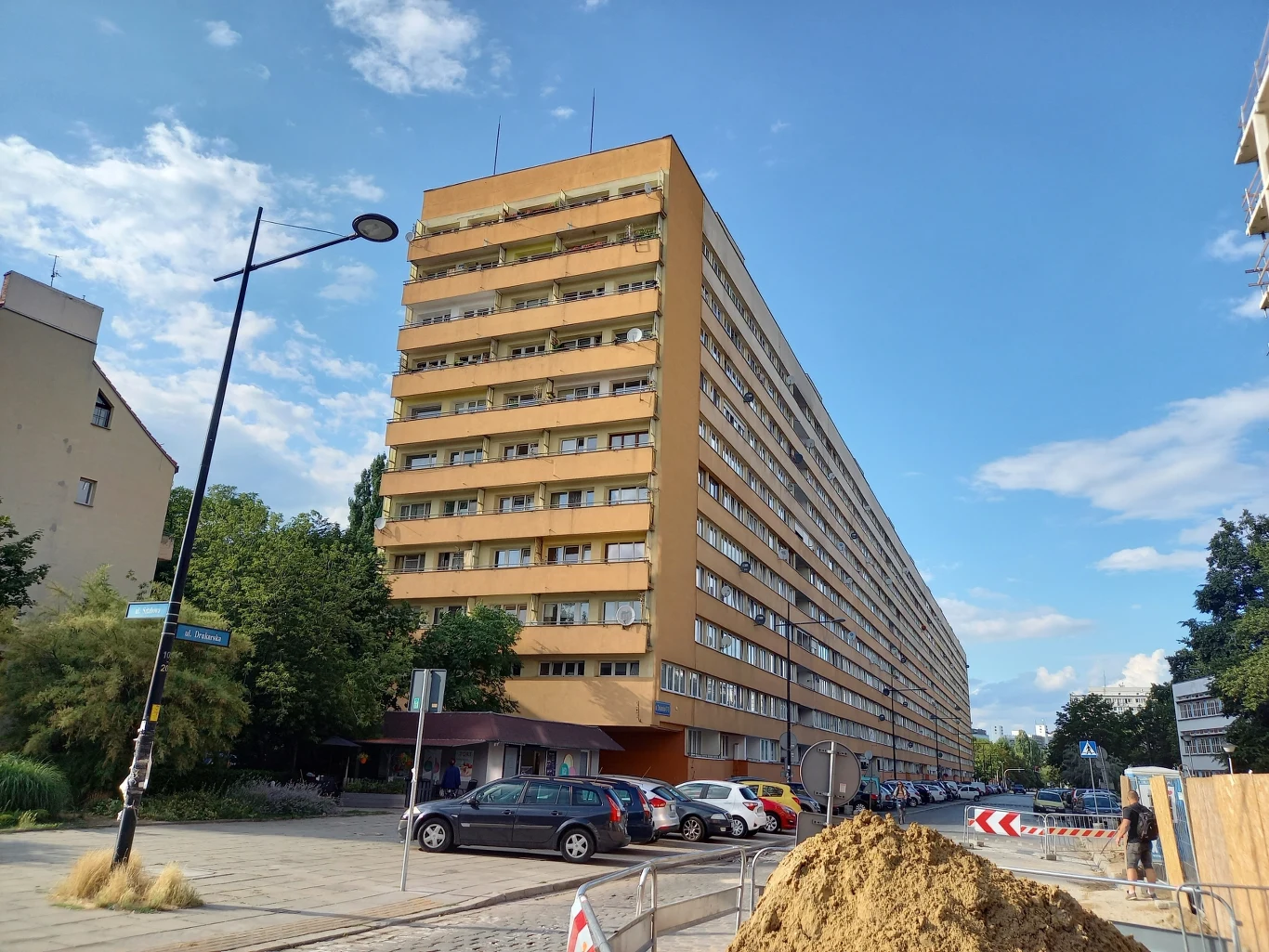 Wysoki blok mieszkalny z balkonami i wieloma piętrami, ustawiony w charakterystycznej perspektywie na tle niebieskiego nieba. Przed budynkiem zaparkowane są samochody, obok znajduje się ulica i plac budowy z kopcem ziemi i elementami zabezpieczającymi ...