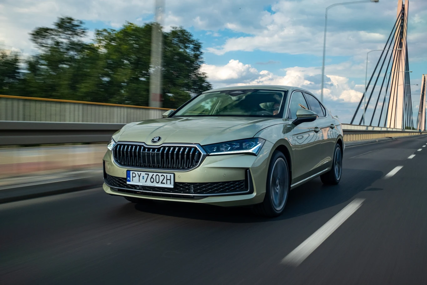 Jasnozielony sedan Skoda jedzie mostem z wyraźnymi liniami jezdni, w tle nowoczesne przęsła mostu i drzewa, na niebie pojawiają się chmury.