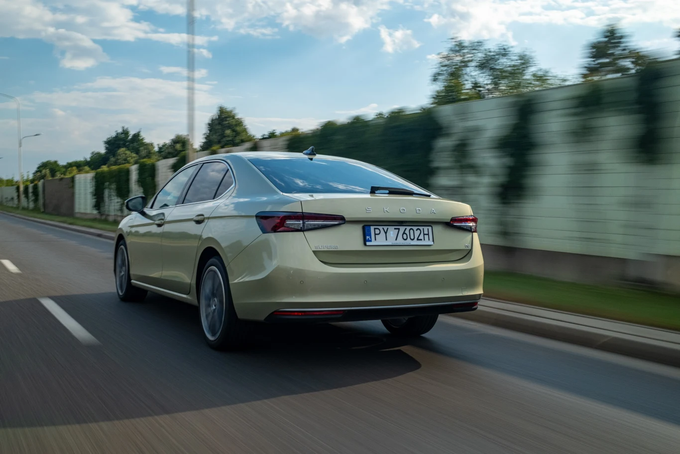 Jasnozielony samochód osobowy marki Skoda jedzie po asfaltowej drodze obok ogrodzenia, w tle widoczne drzewa i niebo z lekkimi chmurami.