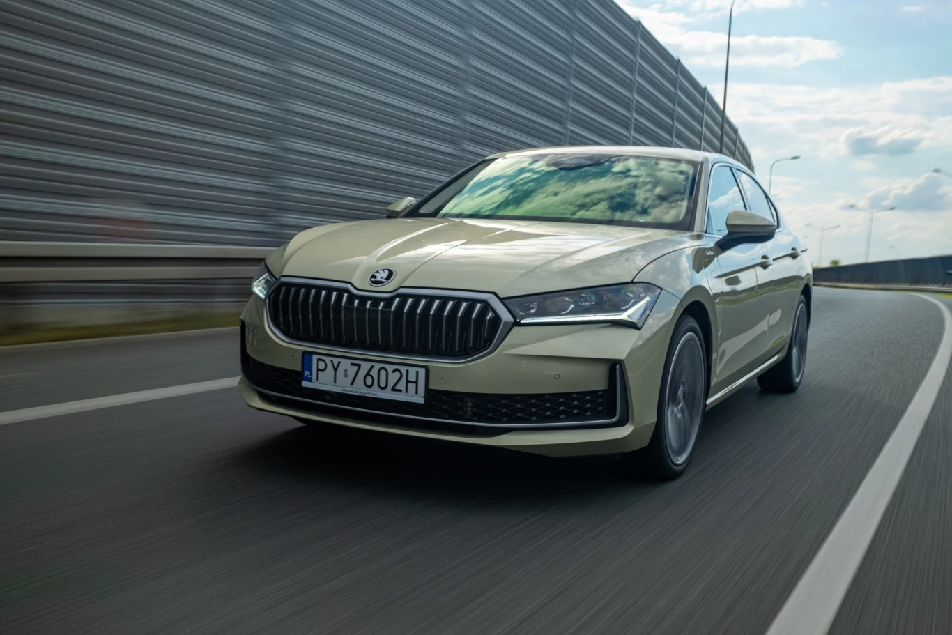 Jasnozielony samochód osobowy marki Škoda jedzie drogą szybkiego ruchu, otoczony barierkami dźwiękochłonnymi, na tle niebieskiego nieba z chmurami.