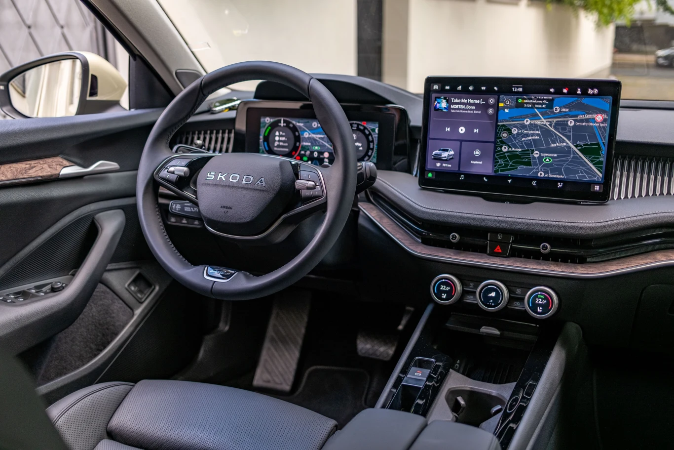 Kokpit nowoczesnego samochodu marki Škoda z dużym ekranem dotykowym na środku deski rozdzielczej, cyfrowym wyświetlaczem za kierownicą oraz trzema pokrętłami do sterowania klimatyzacją; wnętrze jest czyste i nowoczesne z elementami czarnych materiałów ...