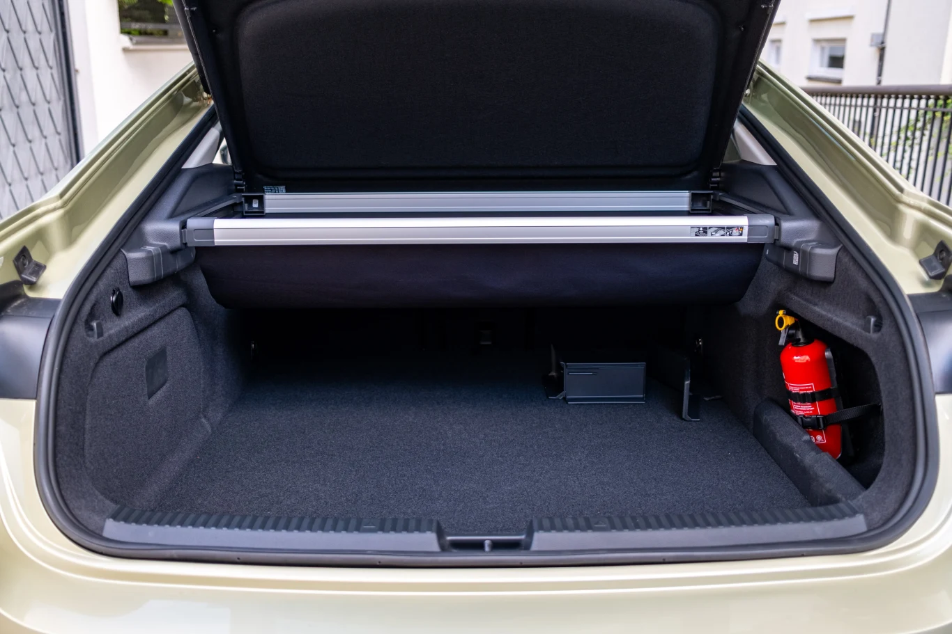 Otwarty bagażnik samochodu osobowego z podniesioną roletą, po prawej stronie znajduje się czerwona gaśnica, wnętrze bagażnika jest czyste i dobrze zorganizowane.