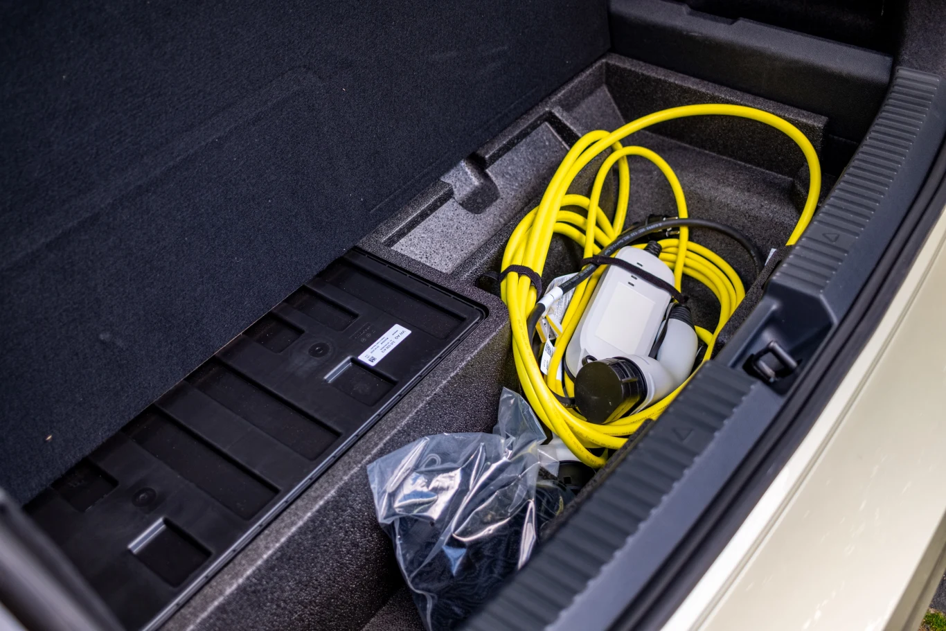 Przestrzeń bagażnika samochodu z wyraźnie widocznym żółtym kablem do ładowania samochodu elektrycznego oraz plastikową torbą z akcesoriami.