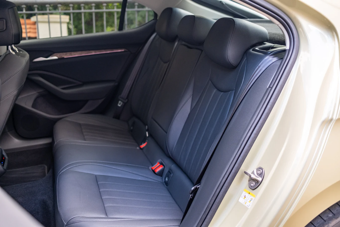 Tylny rząd skórzanych siedzeń samochodu osobowego z trzema zagłówkami i pasami bezpieczeństwa, widoczna otwarta tylna drzwi oraz fragment poręczy ogrodzenia za oknem.