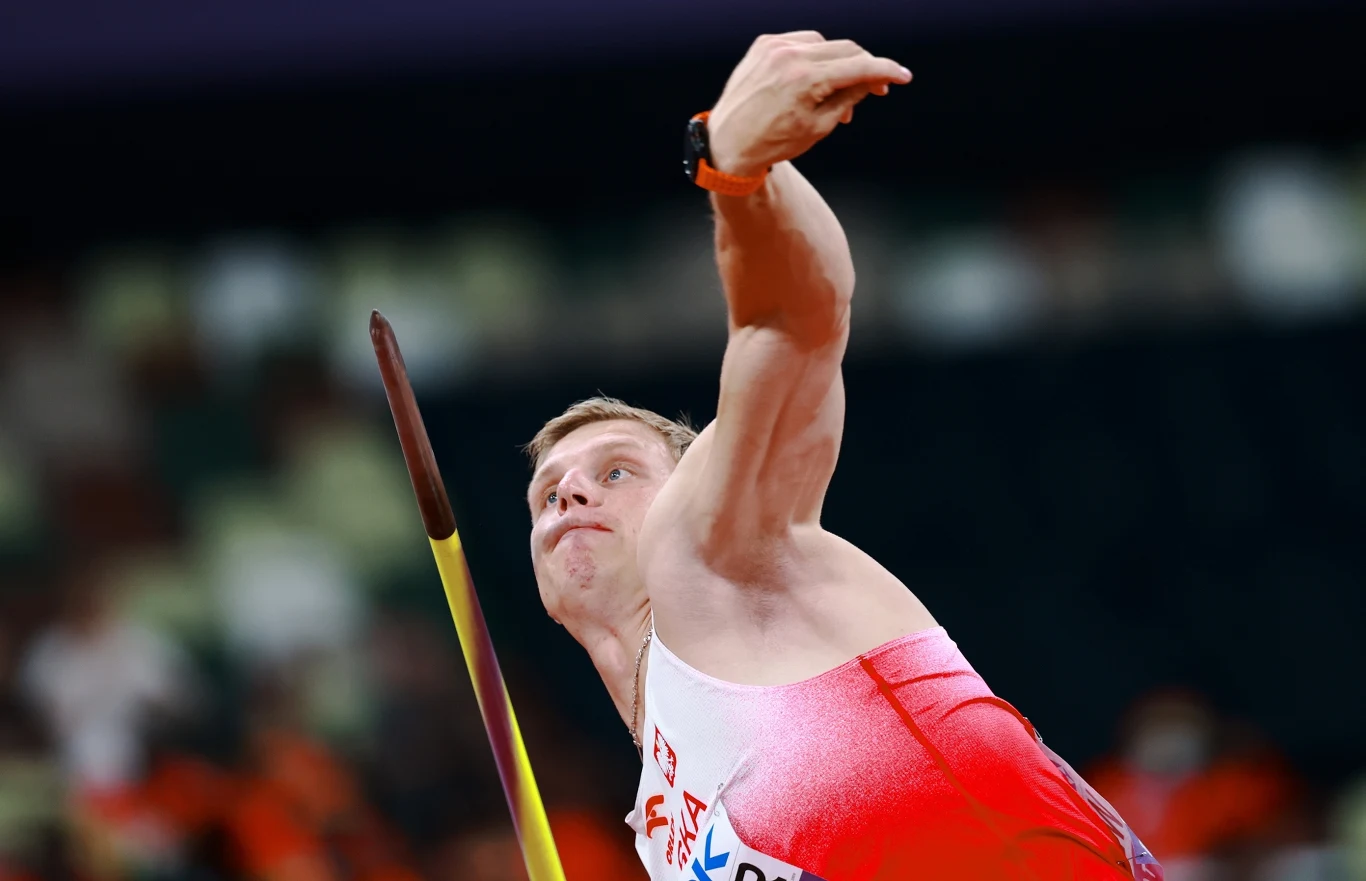 Atleta w białoczerwonym stroju sportowym wykonuje rzut oszczepem na profesjonalnym stadionie, skupiony wyraz twarzy i dynamiczna pozycja ciała podkreślają siłę i precyzję ruchu.