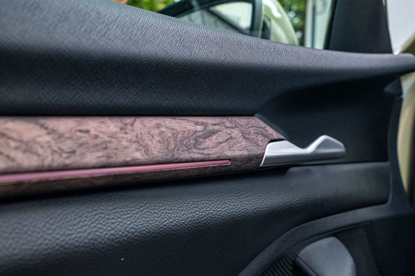 Srebrna klamka oraz drewniany panel dekoracyjny umieszczone na czarnej tapicerce drzwi samochodu, w tle widoczne fragmenty wnętrza auta i rozmyte okno.