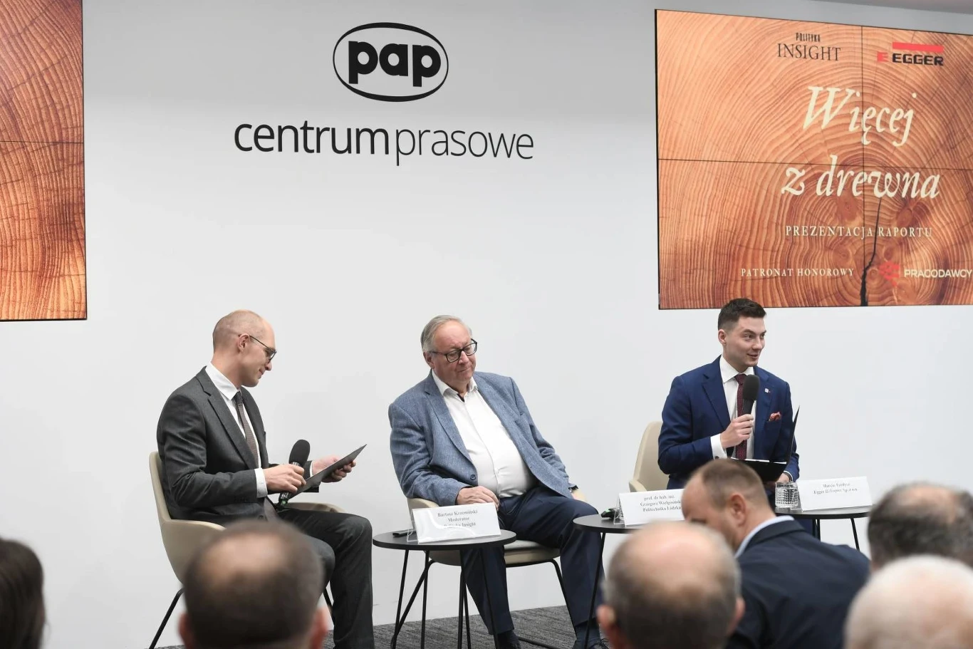 Trzech mężczyzn bierze udział w panelu dyskusyjnym w centrum prasowym, siedzą na krzesłach z mikrofonami, przed nimi widownia, w tle logo PAP oraz prezentacja na temat wykorzystania drewna.