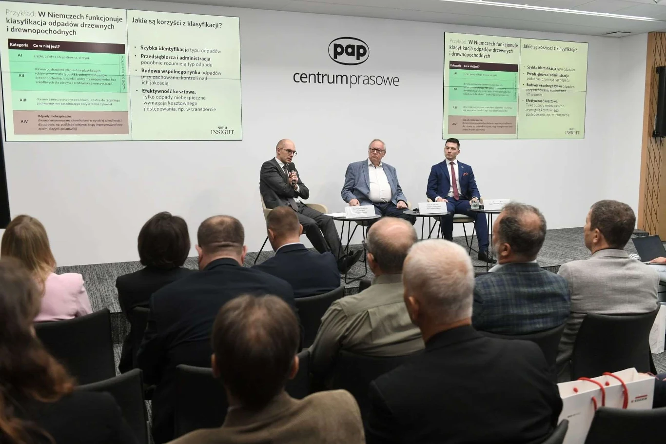 Trzech mężczyzn siedzi na scenie w sali konferencyjnej, za nimi wyświetlone są slajdy prezentacyjne dotyczące klasyfikacji odpadów drzewnych, na widowni zasiada kilku uczestników.