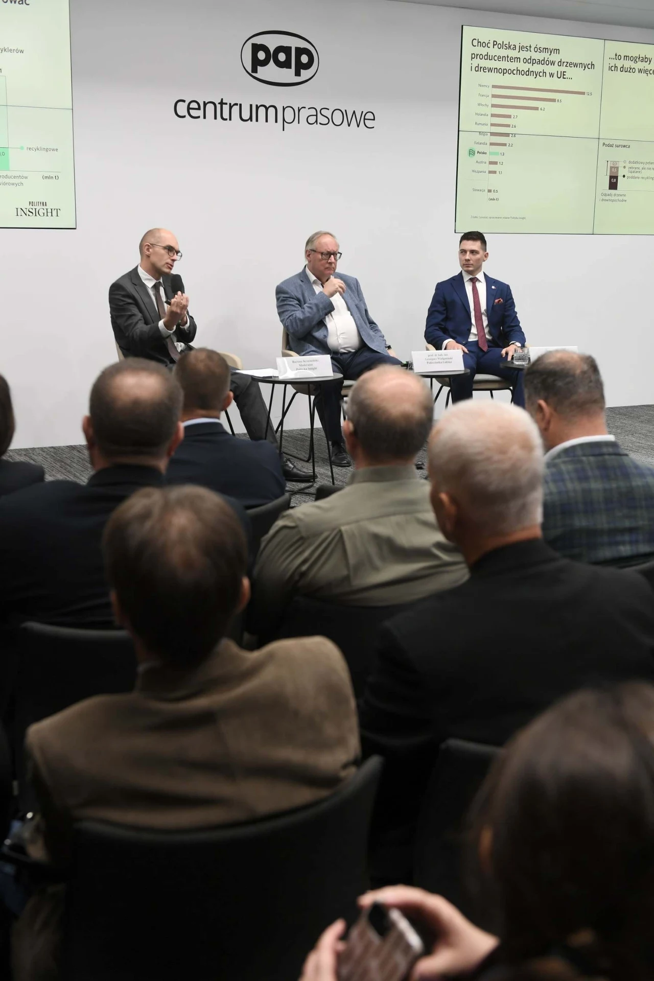Trzech mężczyzn prowadzi panel dyskusyjny na scenie przed publicznością, w tle ekrany z prezentacją, na ścianie logo centrum prasowego PAP.