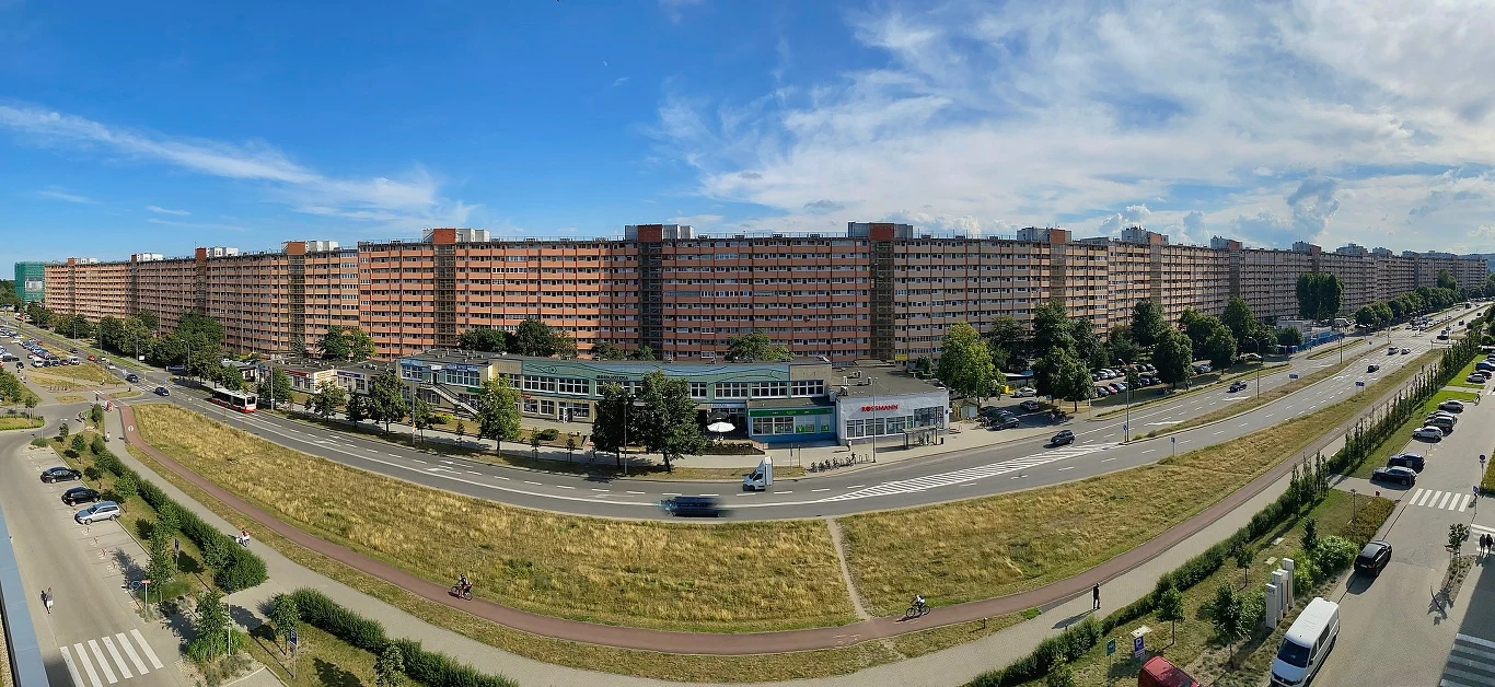 Ogromny blok mieszkalny o charakterystycznej, ciągnącej się na długość zabudowie, przed którym biegnie szeroka ulica z samochodami oraz chodnikiem i ścieżką rowerową, uzupełniona o niewielkie punkty usługowe oraz tereny zielone.