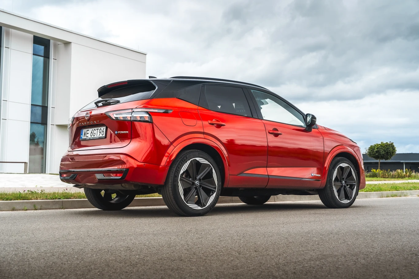 Czerwony nowoczesny samochód typu SUV zaparkowany na ulicy przy nowoczesnym budynku, widok od tyłu, czarne elementy dachu i felg kontrastujące z karoserią.