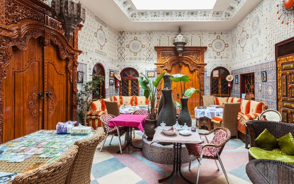 Popularnym noclegiem w Maroko są riady Luksusowy salon ozdobiony bogatymi, drewnianymi drzwiami, orientalnymi mozaikami na ścianach, kolorowymi poduszkami na kanapach oraz ceramicznymi i wiklinowymi stolikami, na których stoją duże wazony z liśćmi roślin.