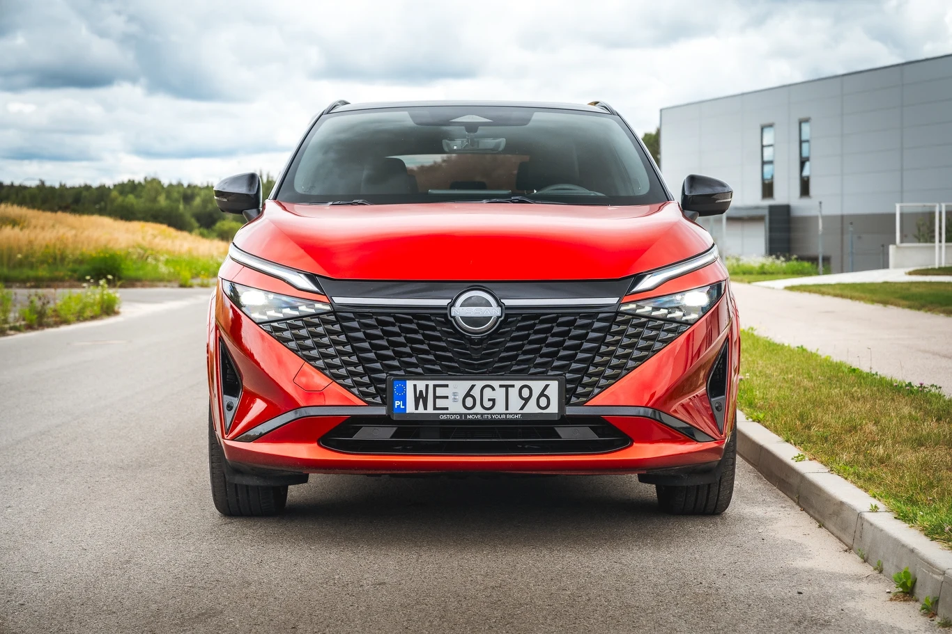 Czerwony nowoczesny samochód osobowy marki Nissan stoi zaparkowany na ulicy w pobliżu budynku o nowoczesnej architekturze, w tle widoczne są drzewa oraz niebo z lekkim zachmurzeniem.
