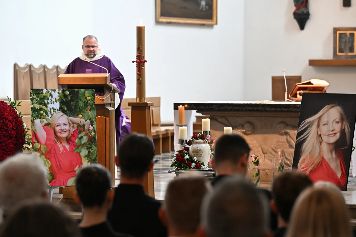 Wnętrze kościoła podczas ceremonii pogrzebowej, za ołtarzem ksiądz wygłasza mowę, po obu stronach ołtarza znajdują się duże portrety uśmiechniętej kobiety w czerwonej sukience, przed ołtarzem liczne kwiaty i znicze, a w ławkach siedzą uczestnicy uroczy...