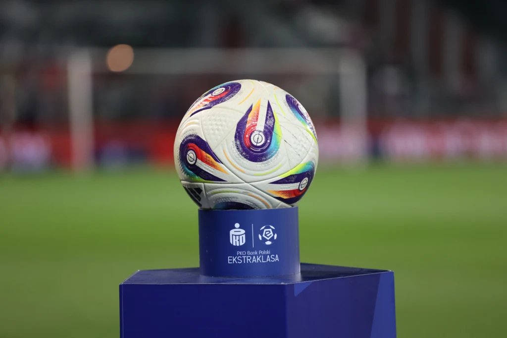 Piłka do piłki nożnej ustawiona na niebieskim postumencie z logotypami Polskiej Ekstraklasy na tle rozmytego stadionu piłkarskiego.