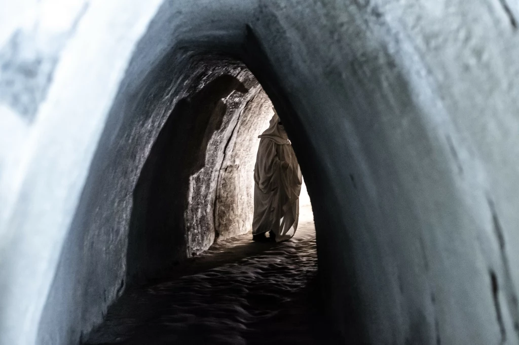Ciemny tunel skalny z osobą w białym stroju idącą w kierunku światła na końcu korytarza, wywołujący efekt tajemniczości i skupienia na postaci.