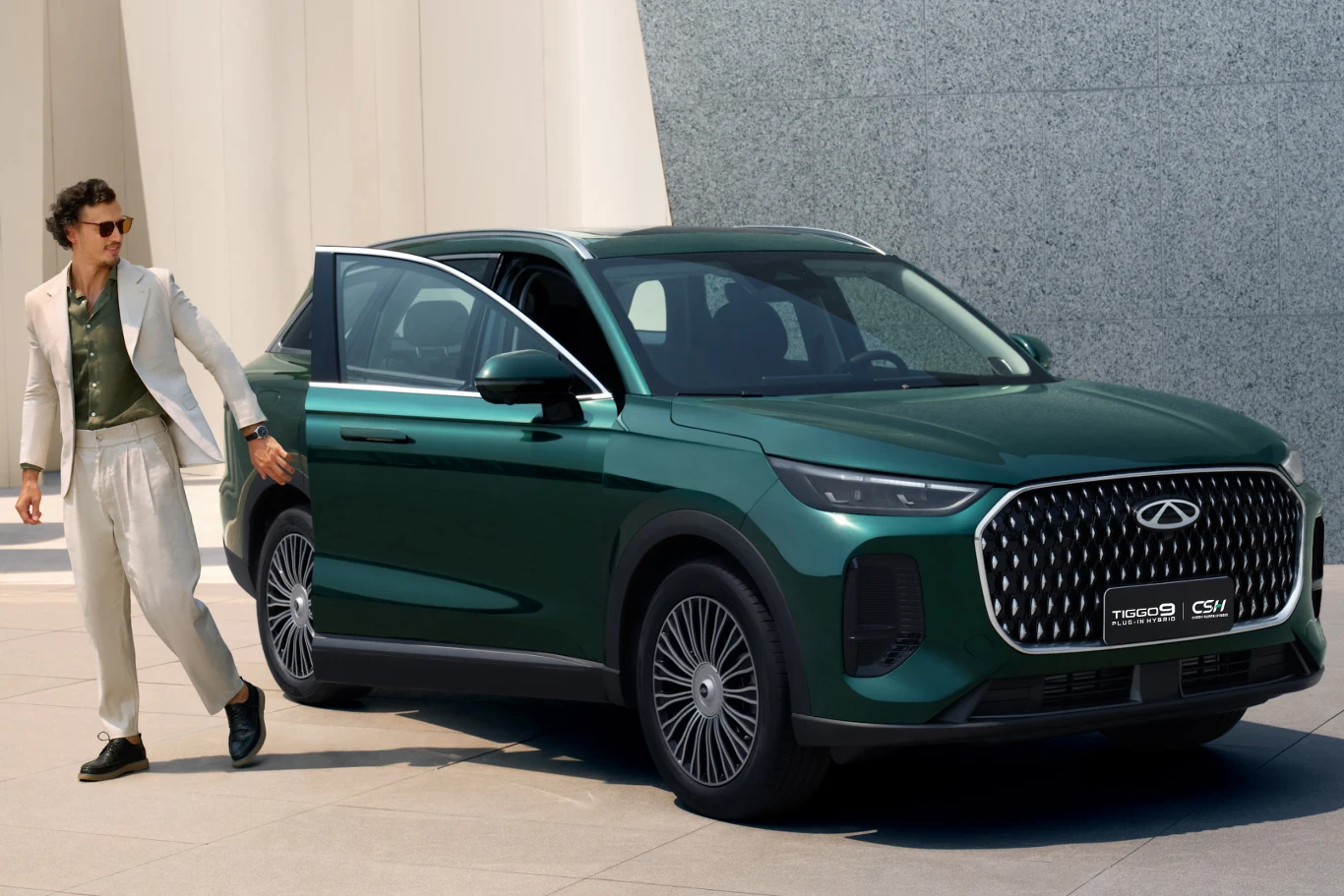 Tiggo 9 PHEV to jeden z dwóch nowych modeli, który trafi do sprzedaży w Polsce. Mężczyzna w jasnym garniturze opuszcza zielony samochód SUV marki Chery stojący przed nowoczesnym budynkiem o minimalistycznej architekturze.