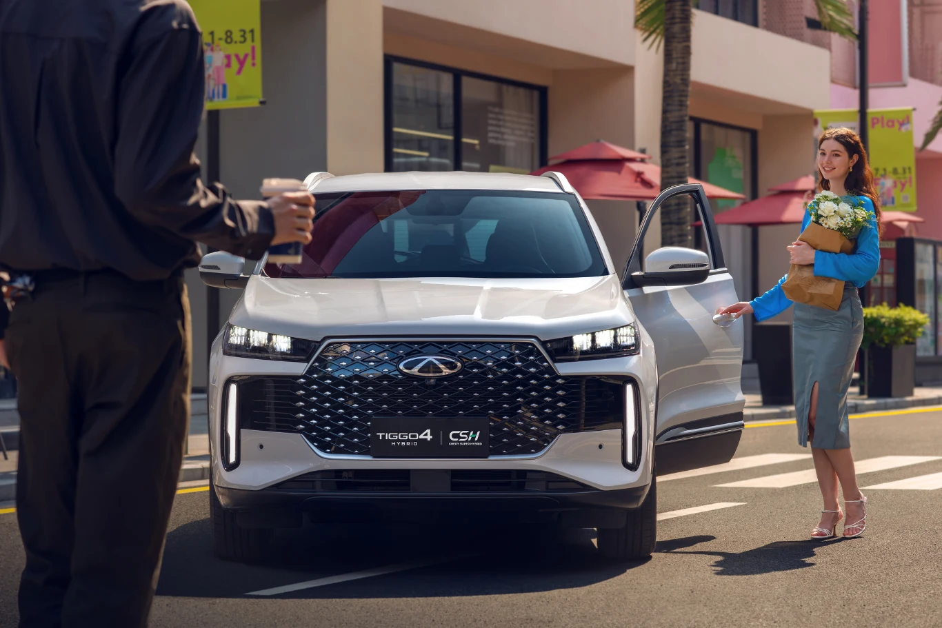 Tiggo 4 HEV to kompaktowy SUV z napędem hybrydowym. Kobieta w eleganckim ubraniu z bukietem białych kwiatów wychodzi z białego SUV-a Chery Tiggo 4 na tle miejskiej ulicy, po lewej stronie widoczny mężczyzna trzymający kubek z kawą.