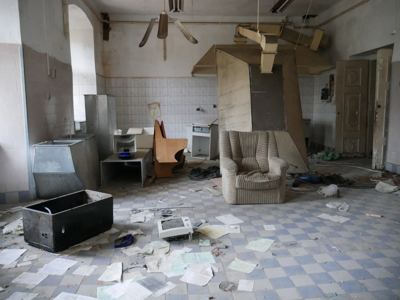 Zdemolowane pomieszczenie z przewróconymi meblami, rozrzuconymi papierami na podłodze i zniszczonym sprzętem; w centrum znajduje się stara, zużyta kanapa, a na ścianach widać zabrudzenia i ślady zaniedbania.