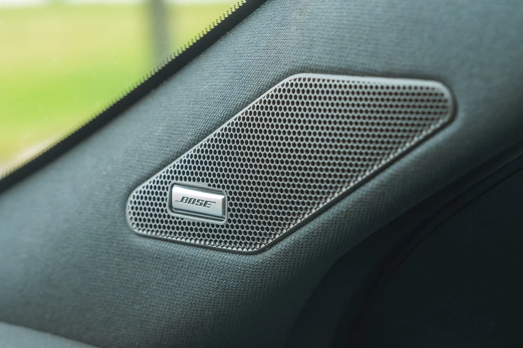 Nagłośnienie Bose kosztuje 3 tys. zł i obejmuje 8 głośników, wzmacniacz i subwoofer Srebrny głośnik marki Bose wmontowany w tapicerkę samochodową w pobliżu szyby, otoczony czarnym materiałem podsufitki.