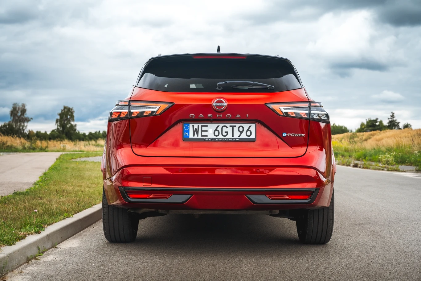 Czerwony samochód marki Nissan Qashqai e-Power widoczny od tyłu, zaparkowany na drodze w otoczeniu zielonych pól i pod pochmurnym niebem.