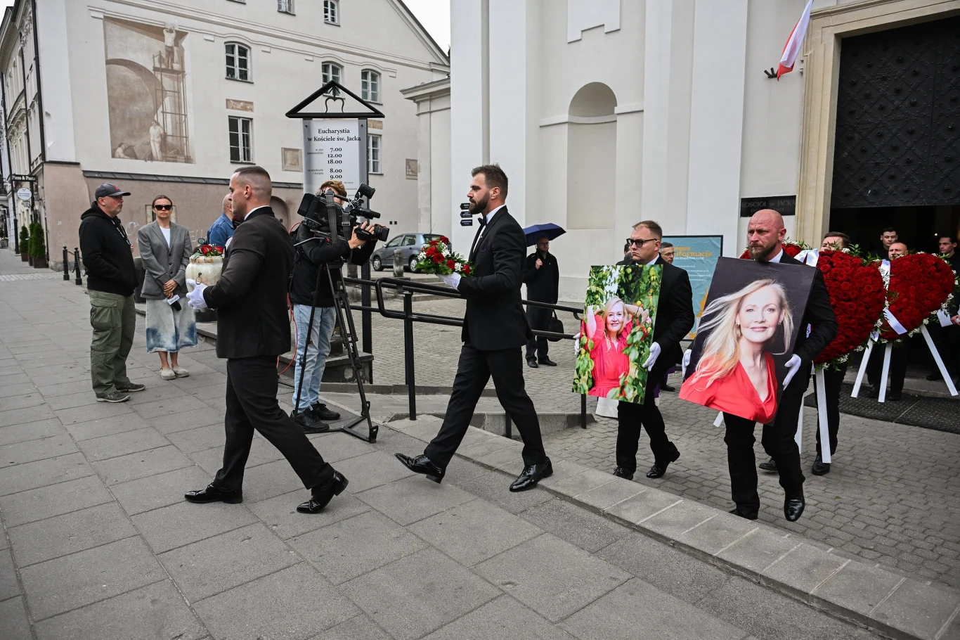 Kondukt żałobny przed wejściem do kościoła, mężczyźni ubrani na czarno niosą wieniec oraz dużą fotografię młodej kobiety, w tle wieńce pogrzebowe i osoby uczestniczące w wydarzeniu.