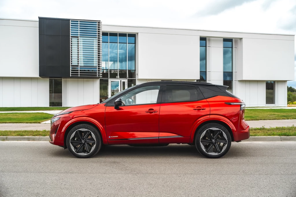 Nissan Qashqai w czerwonym kolorze i wersji N-Design przyciąga wzrok na ulicy Czerwony samochód osobowy stojący na jezdni przed nowoczesnym, białym budynkiem z dużymi przeszkleniami i czarnym panelem dekoracyjnym.