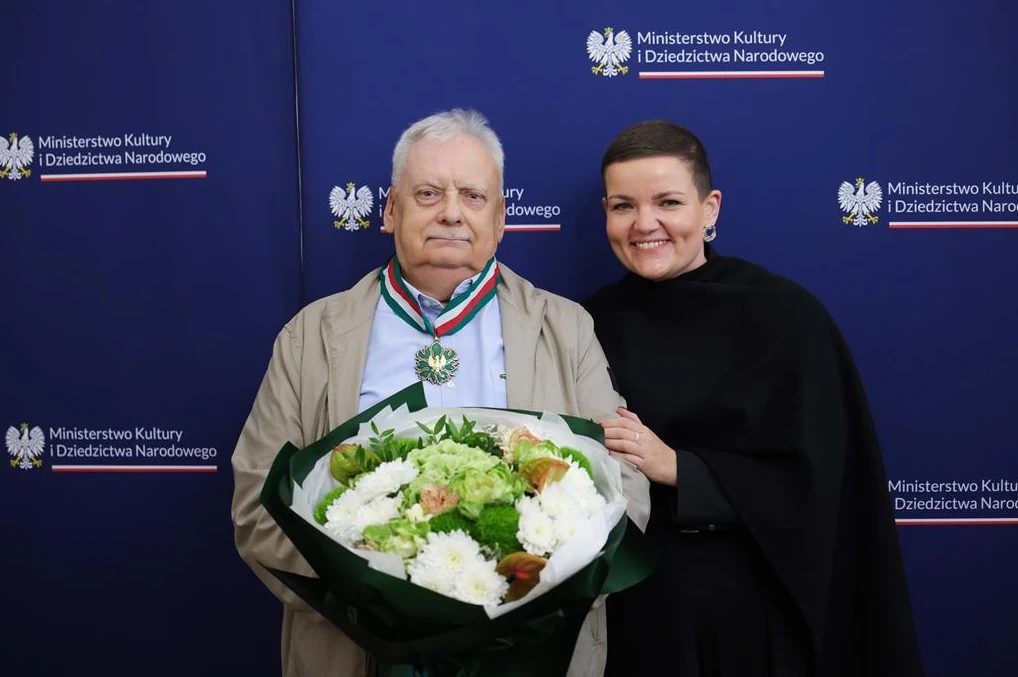 Andrzej Sapkowski i Marta Cienkowska / źródło: gov.pl Starszy mężczyzna z medalem na szyi trzyma w rękach bukiet kwiatów, obok niego stoi uśmiechnięta kobieta w czarnej pelerynie, w tle widoczne logo Ministerstwa Kultury i Dziedzictwa Narodowego na niebieskim tle.
