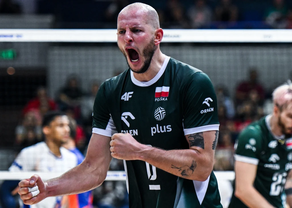 Bartosz Kurek w barwach reprezentacji Polski Siatkarz w ciemnozielonym stroju z emblematami reprezentacji Polski wyraża silne emocje, ściskając pięść i krzycząc, w tle pozostali zawodnicy i trybuny