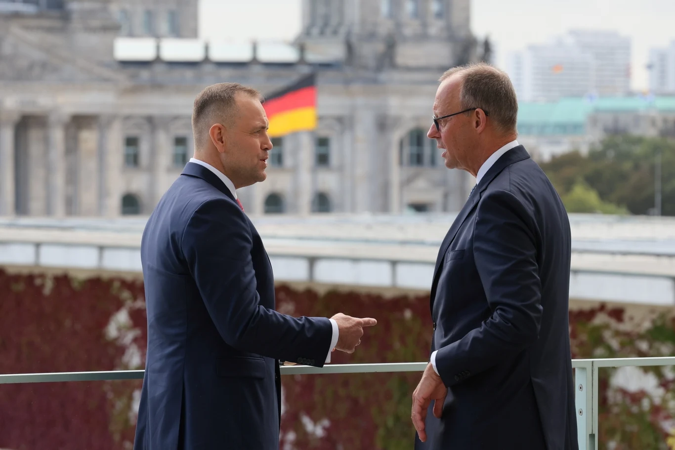 Dwóch mężczyzn w garniturach prowadzi rozmowę na tle budynku z widoczną flagą Niemiec, w tle nowoczesna architektura oraz fragmenty roślinności pokrywającej mury.
