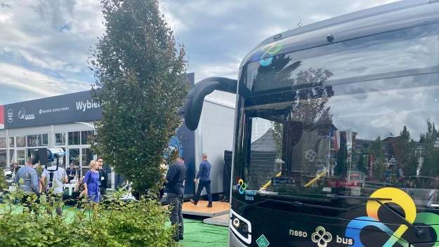 Nowoczesny autobus elektryczny z wyraźnym logo firmy oraz rejestracją, zaparkowany na tle terenów zielonych i pawilonu targowego, wokół znajdują się odwiedzający i uczestnicy wydarzenia.