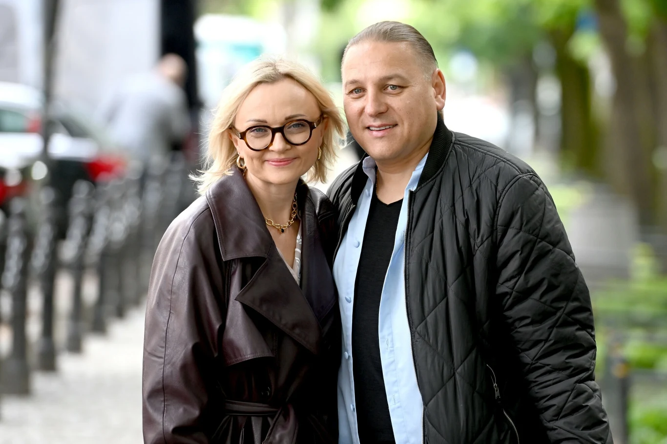 Edyta i Łukasz Golec są małżeństwem od 25 lat Para dorosłych ludzi pozuje razem na tle miejskiej ulicy, kobieta w okularach i brązowym płaszczu, mężczyzna ubrany na jasno z ciemną kurtką, oboje uśmiechnięci, w tle rozmyte drzewa i barierki.