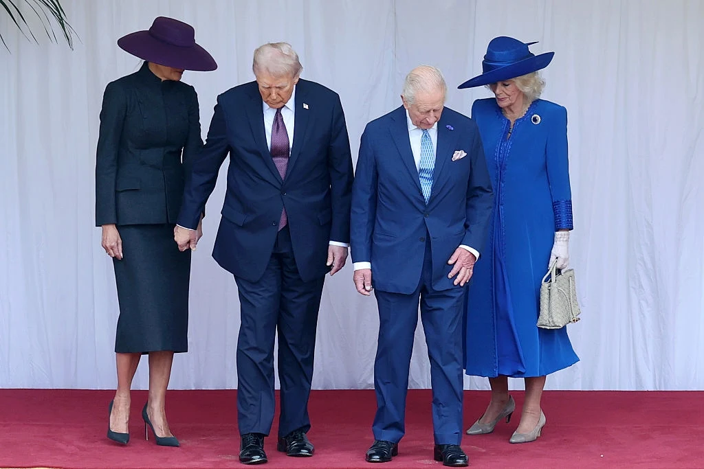 Cztery elegancko ubrane osoby stoją na czerwonym dywanie przed jasnym tłem, dwie kobiety mają na sobie kapelusze i sukienki, dwaj mężczyźni w garniturach pochylają głowy obserwując podłoże.