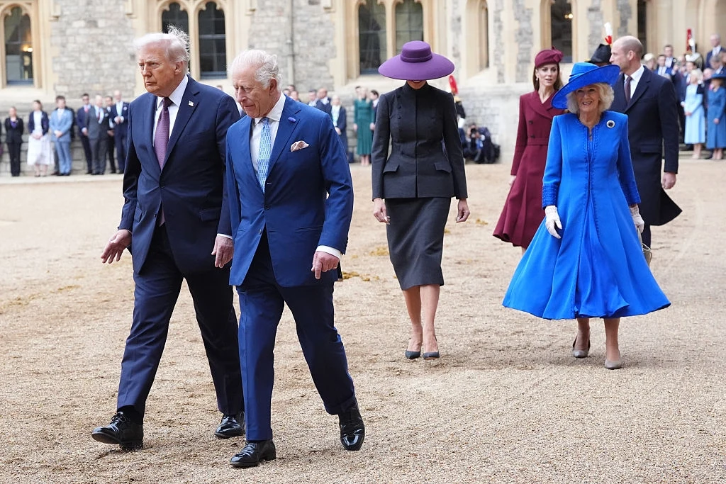 Wizyta pary prezydenckiej USA w Wielkiej Brytanii Grupa elegancko ubranych osób spaceruje po dziedzińcu zamkowym, wśród nich mężczyźni w garniturach i kobiety w stylowych kapeluszach oraz kolorowych sukniach, w tle widać historyczną architekturę oraz zgromadzenie ludzi.