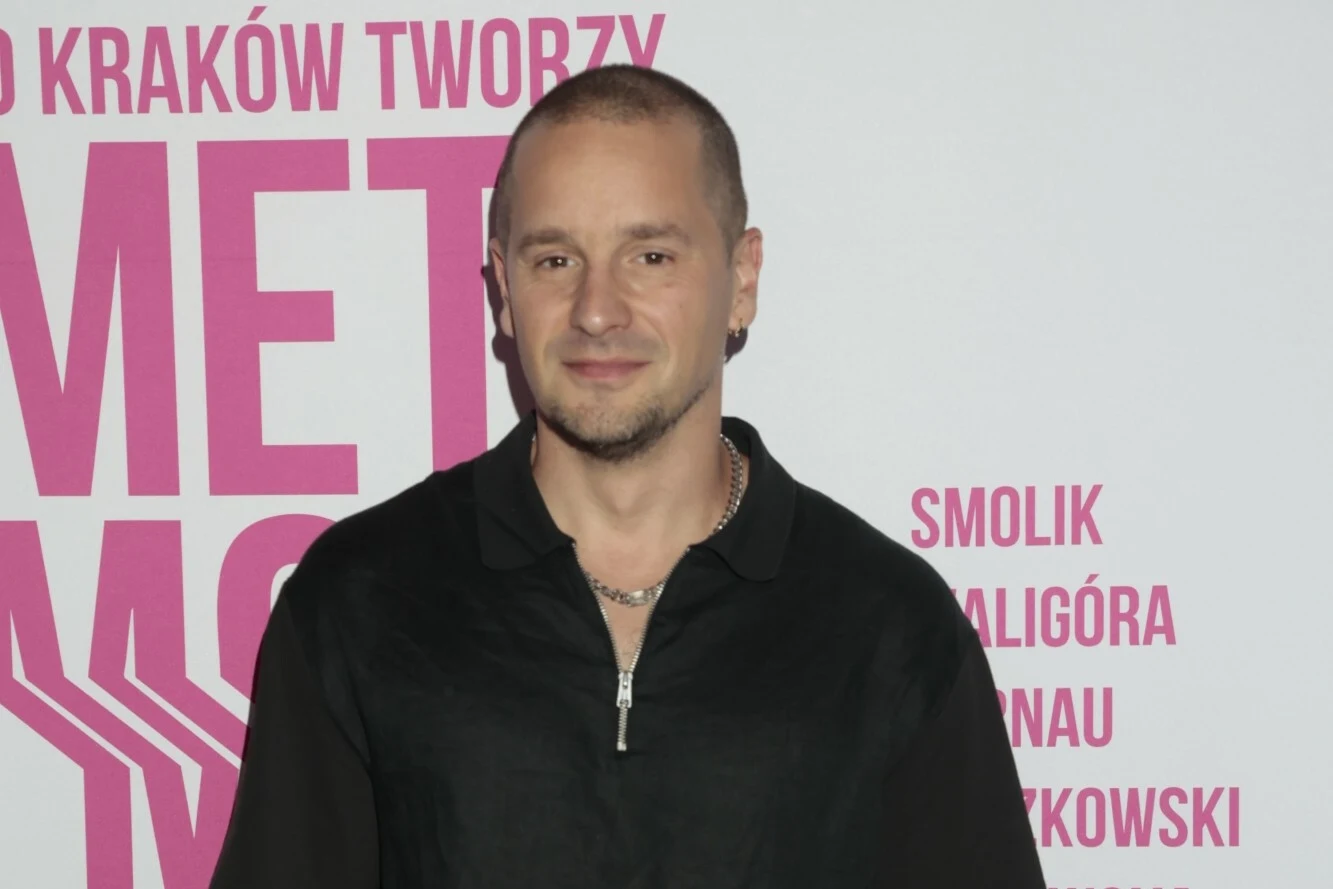 Krzysztof Zalewski podczas konferencji prasowej wydarzenia "To Kraków tworzy metamorfozy" Mężczyzna w czarnej koszuli stoi na tle tablicy z dużymi, różowymi napisami, na której widoczne są nazwiska oraz napisy jasnoróżowe na białym tle.