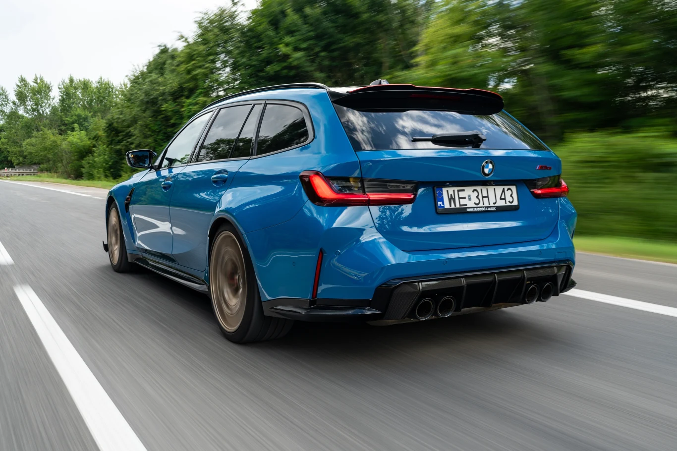 Niebieskie sportowe kombi marki BMW jedzie dynamicznie po asfaltowej drodze, otoczonej przez zieleń i drzewa, widok z tyłu pozwala dostrzec tablicę rejestracyjną oraz charakterystyczne czarne felgi i dyfuzor.