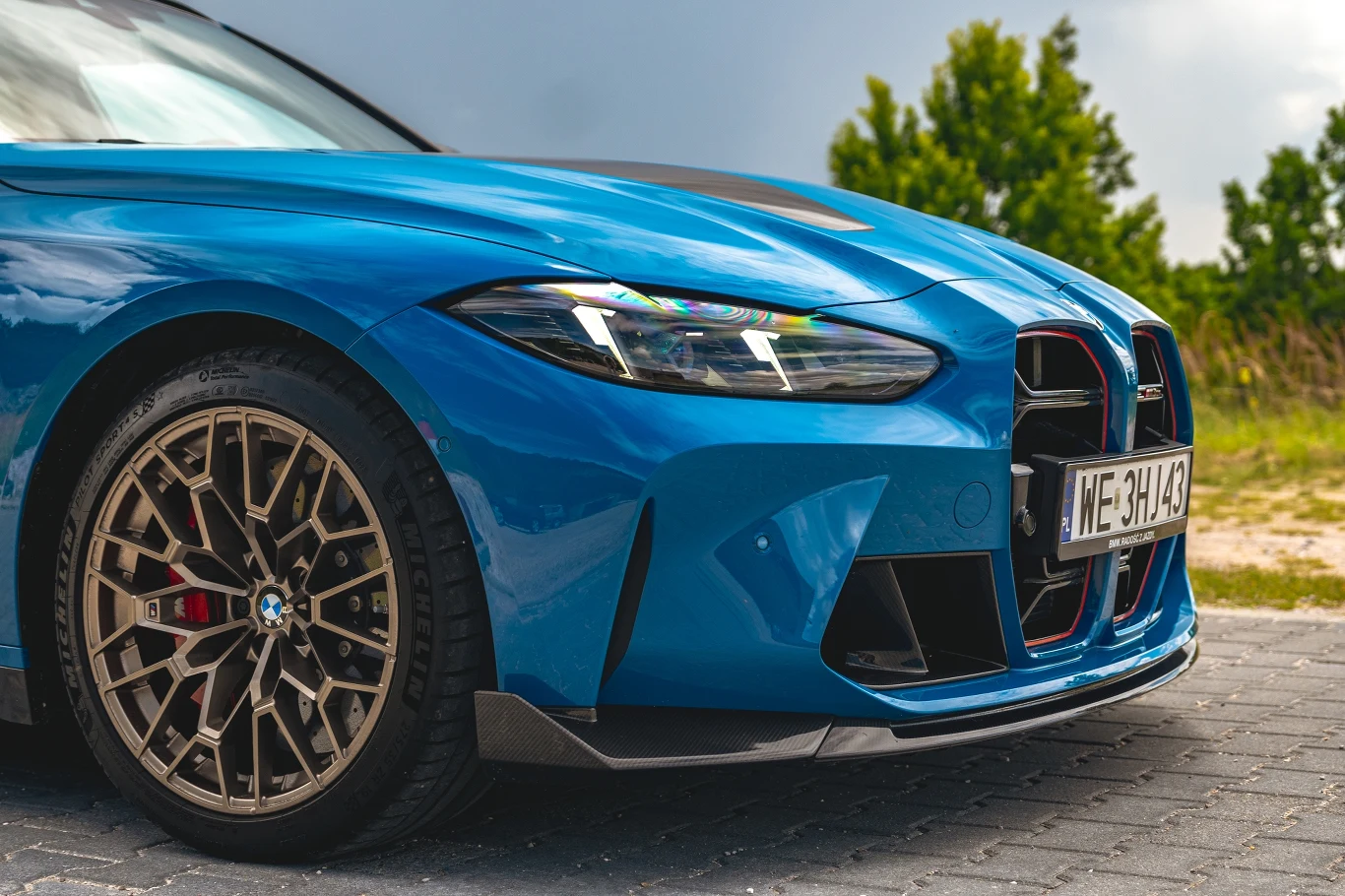 Przednie części niebieskiego sportowego samochodu BMW z wyeksponowaną atrapą chłodnicy, eleganckimi reflektorami i aluminiową felgą na tle zieleni i nieba.