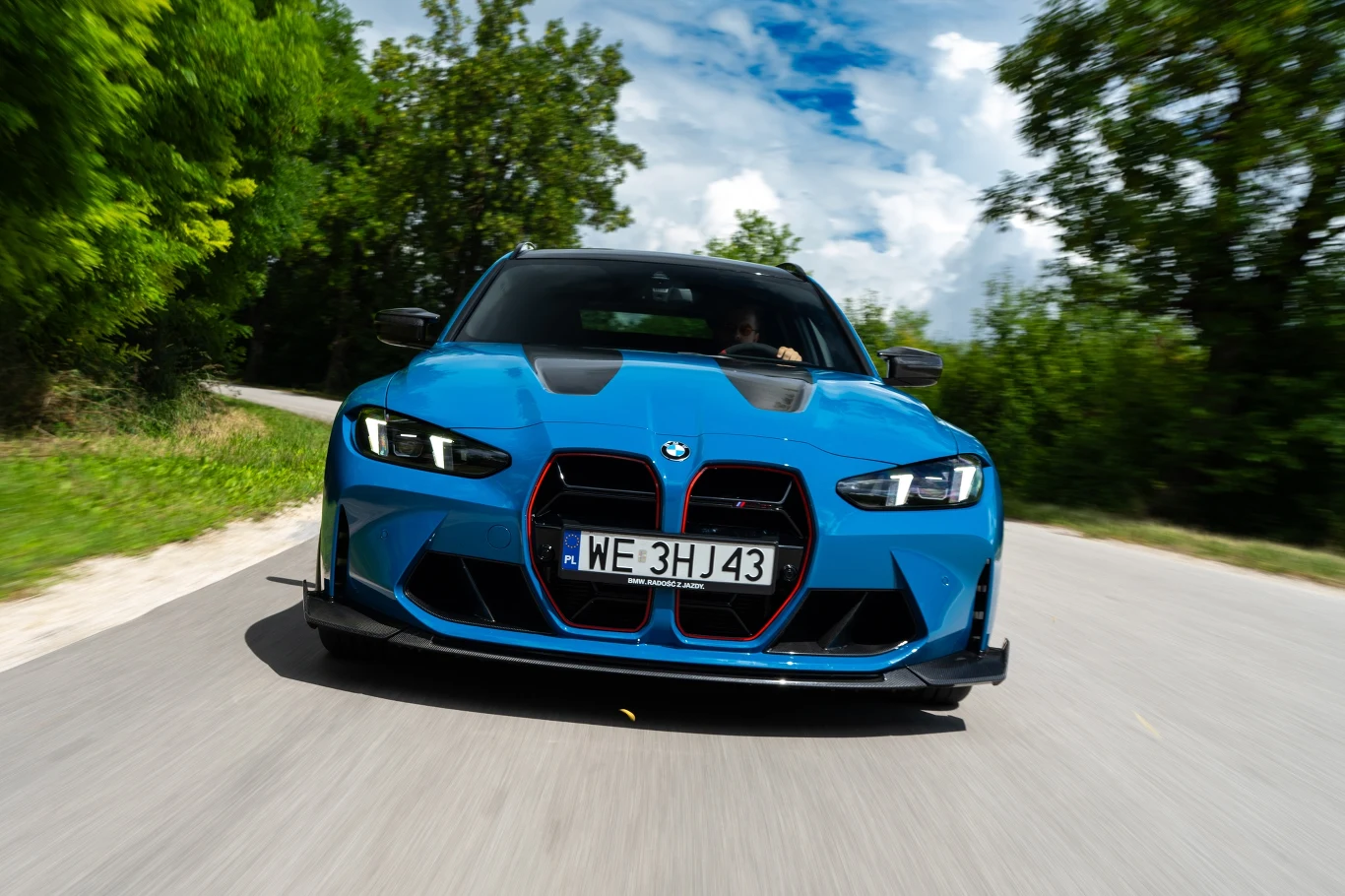 Jasnoniebieskie sportowe BMW z agresywnym grillem i czarnymi akcentami pędzi krętą, asfaltową drogą otoczoną zielonymi drzewami i błękitnym niebem.