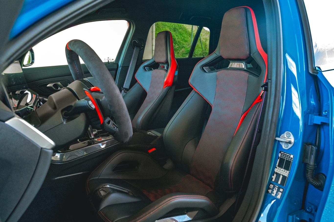 Nowoczesne wnętrze samochodu sportowego z dwoma kubełkowymi fotelami wykończonymi czarną i czerwoną tapicerką, kierownicą sportową oraz detalami ze skóry i alcantary, widoczne przez otwarte drzwi pojazdu.